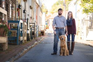 A couple in a portrait standing on a downtown street with their dog.