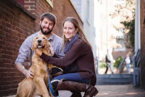 A couple poses with their dog in front of a brick building for an engagement photo in downtown Annapolis.