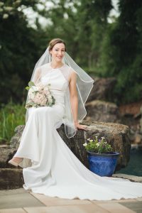 A portrait of a bride in a wedding dress sitting on a rock next to a pool.