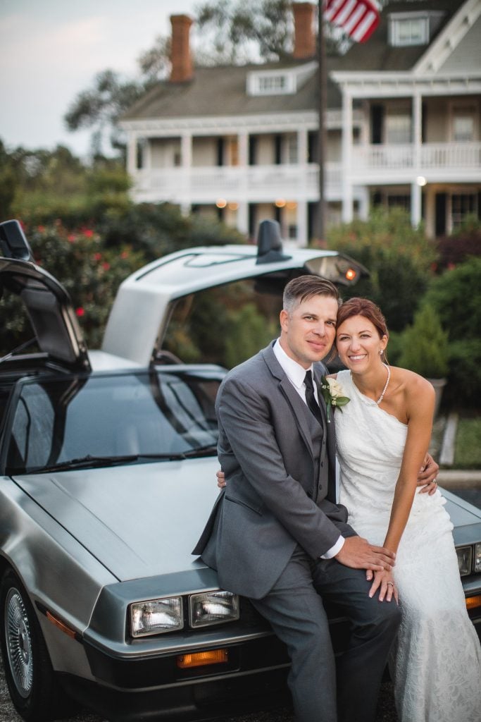 A bride and groom pose next to a Delorean car on their wedding day.