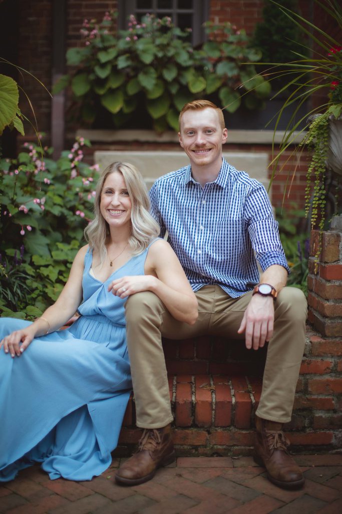 An engaged couple sitting on bricks in Downtown Annapolis, capturing a beautiful engagement photo in a garden.