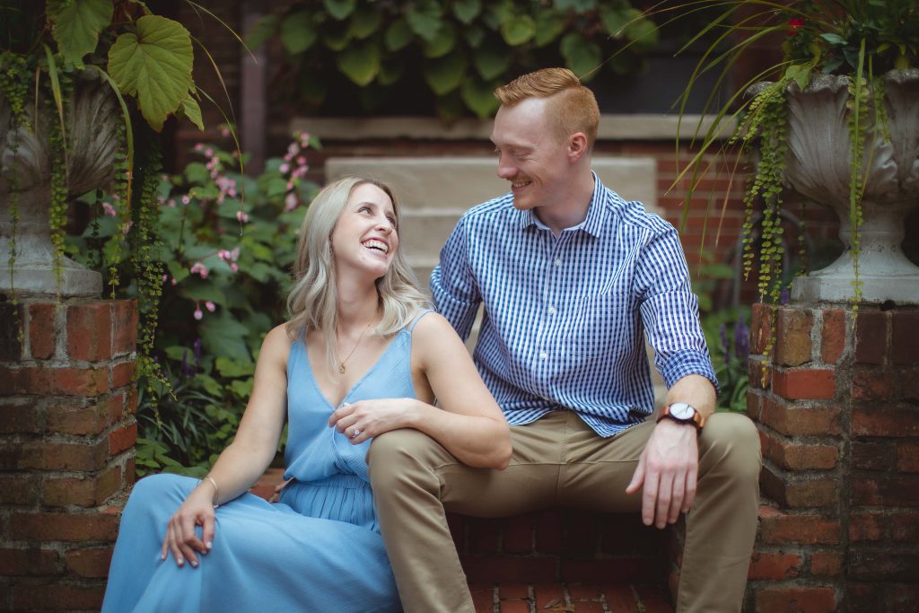 An engaged couple sitting on bricks in a garden for their Downtown Annapolis engagement portrait.