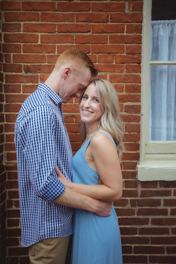An engaged couple embracing in front of a brick wall in downtown Annapolis, capturing a beautiful engagement photo portrait.