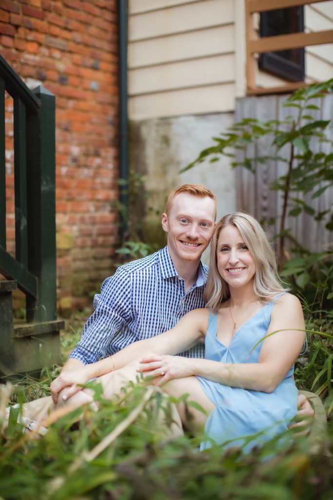 An engaged couple having a portrait taken in Downtown Annapolis, sitting in the grass in front of a brick building.