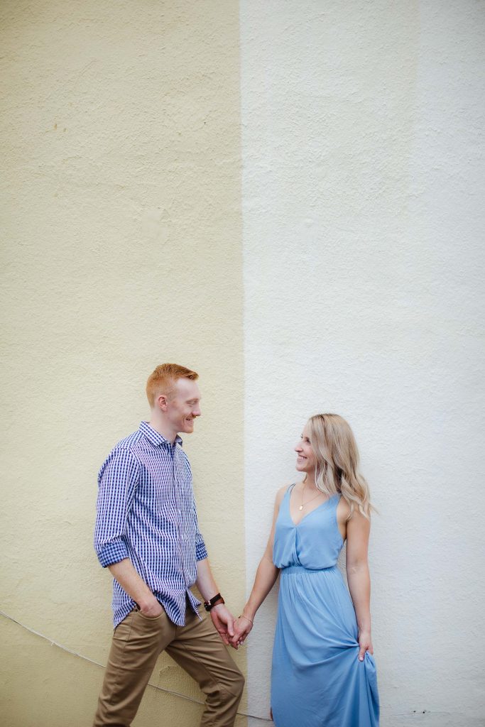 An engaged couple in Downtown Annapolis, holding hands in front of a yellow wall for their engagement photo portrait.