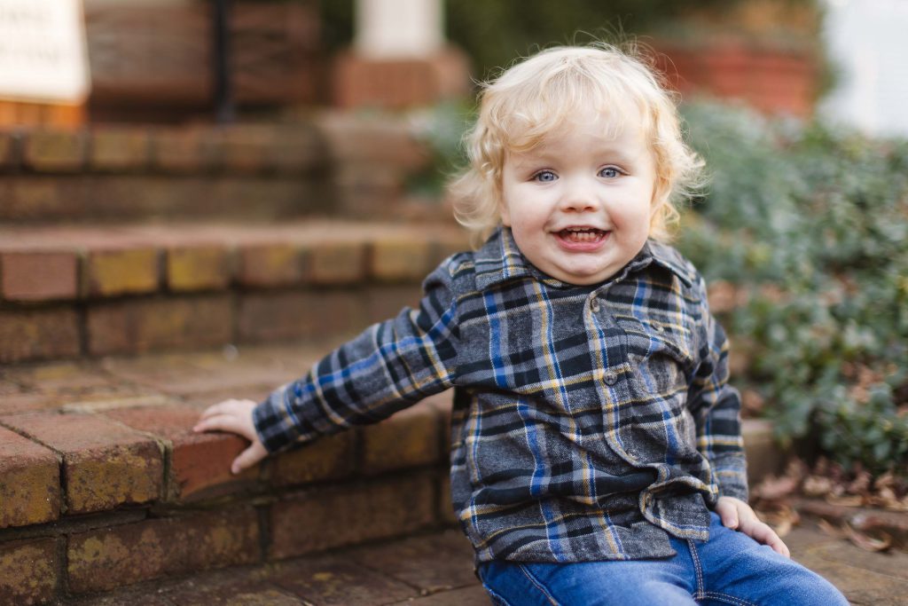 A toddler sits on the steps of a house during a family engagement session.