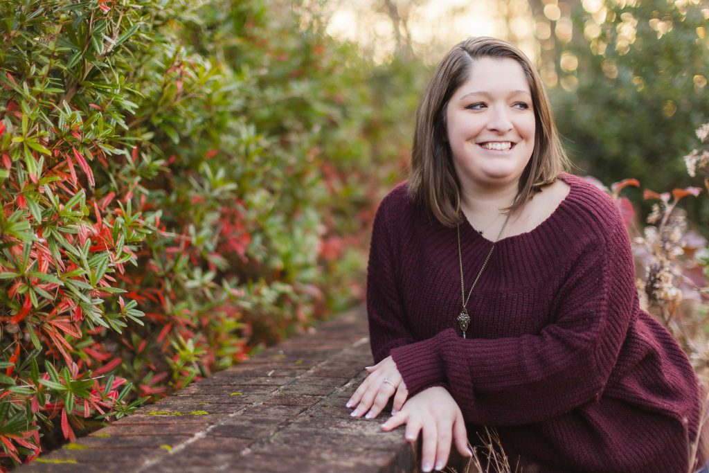 A woman in a burgundy sweater posing for a portrait against a brick wall at Quiet Waters Park.
