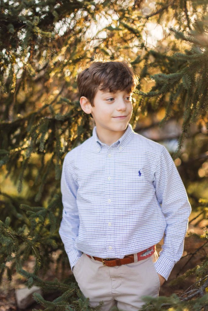 A boy in a blue shirt is posing for a portrait in front of a pine tree.