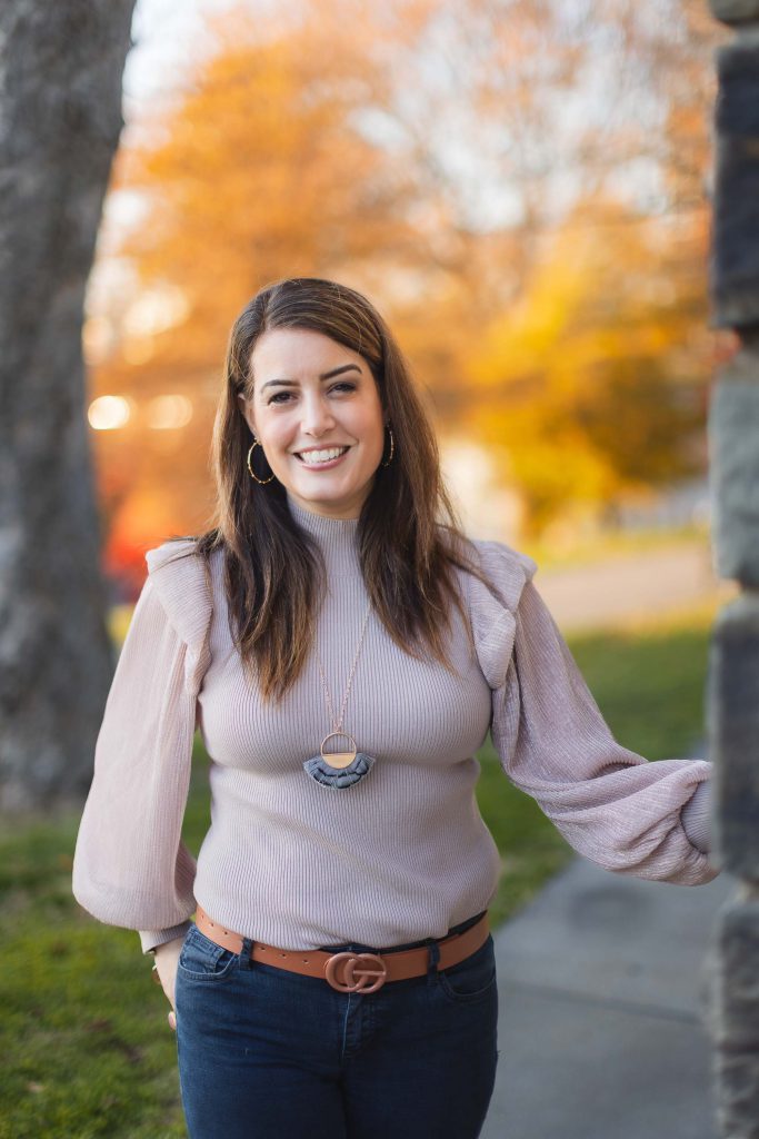 A woman in jeans and a pink top is smiling in front of a brick wall, showcasing her joyful portrait.