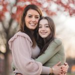 A mother and daughter sharing a warm embrace in front of a vibrant red tree at Federal Hill Park.