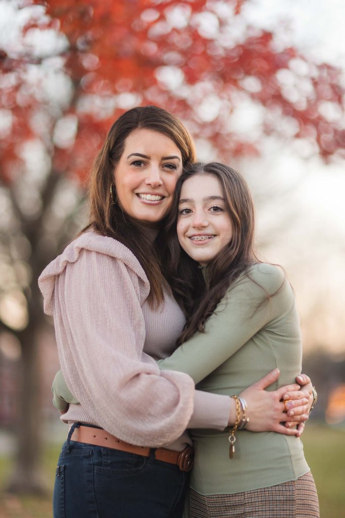 A mother and daughter sharing a warm embrace in front of a vibrant red tree at Federal Hill Park.