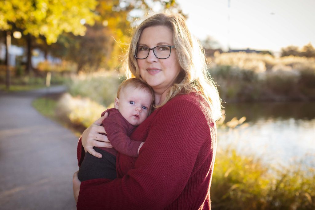 A woman holding a baby in front of a sunset-filled pond.