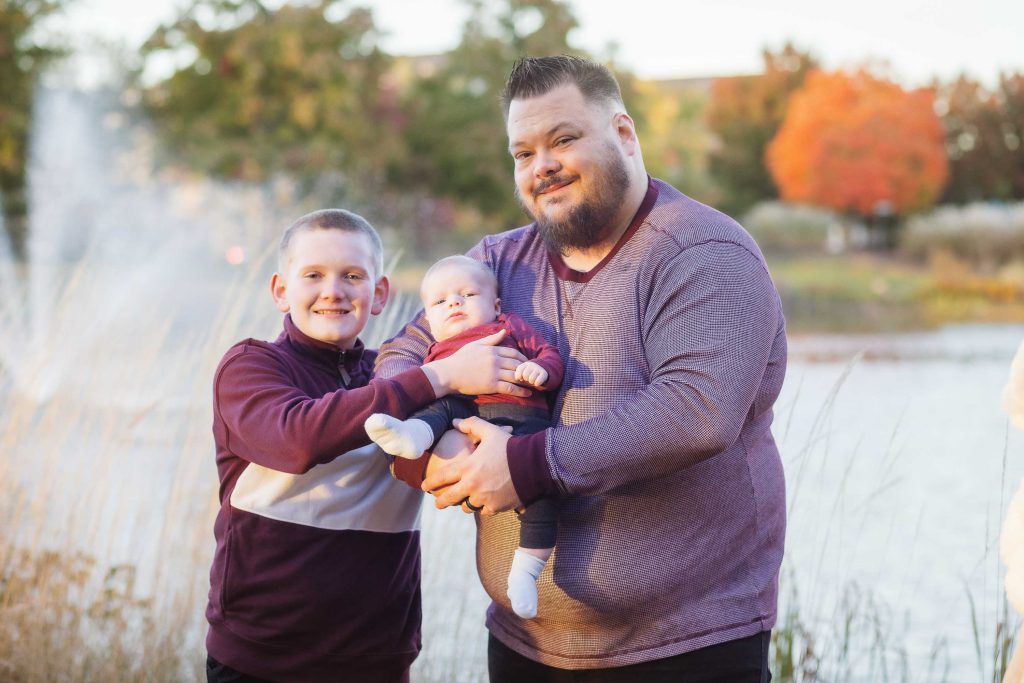 A family portrait of two men holding a baby in front of a serene pond at sunset.