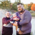 A family portrait of two men holding a baby in front of a serene pond at sunset.