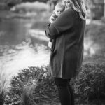 A black and white photo of a woman holding a baby in front of a pond at sunset.