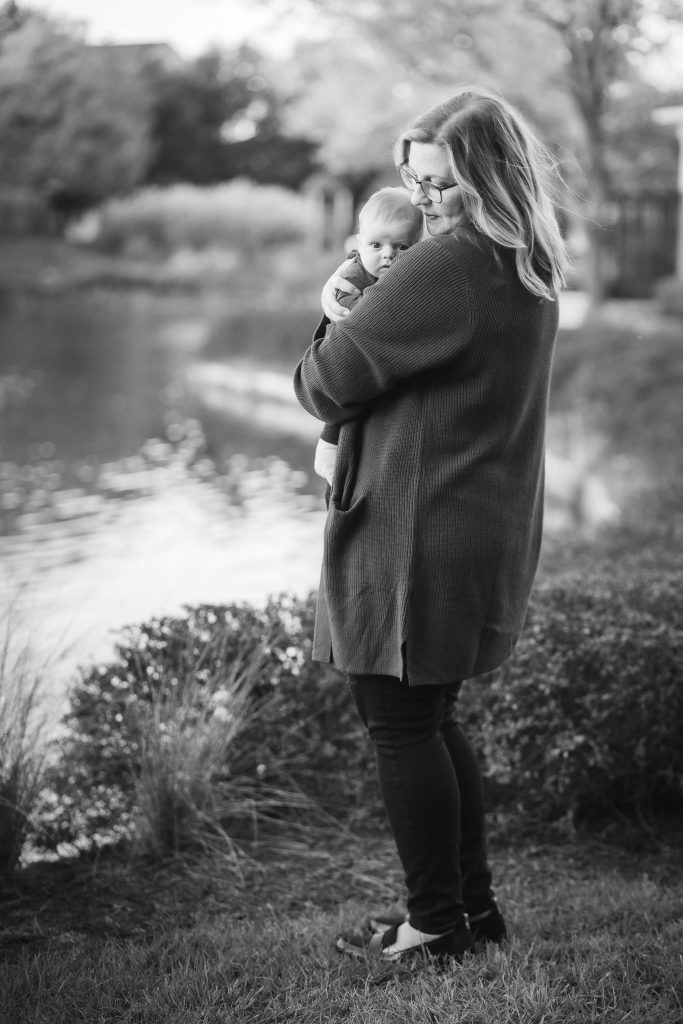 A black and white photo of a woman holding a baby in front of a pond at sunset.