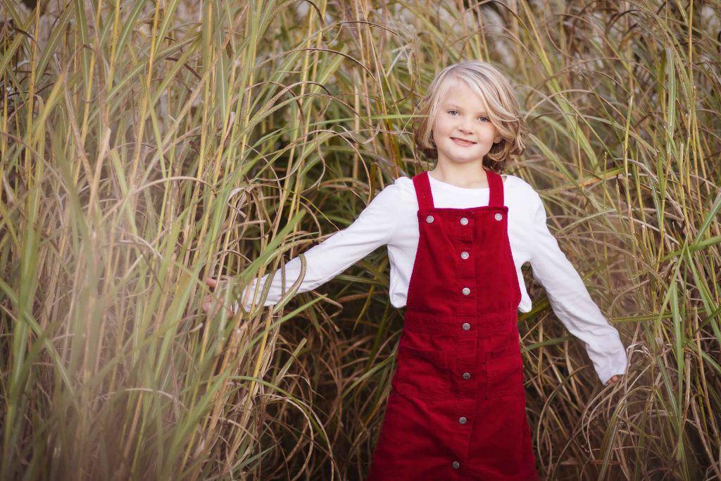A little girl in red overalls standing in tall grass, captured beautifully in a family portrait at sunset.