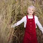 A little girl in red overalls standing in tall grass, captured beautifully in a family portrait at sunset.