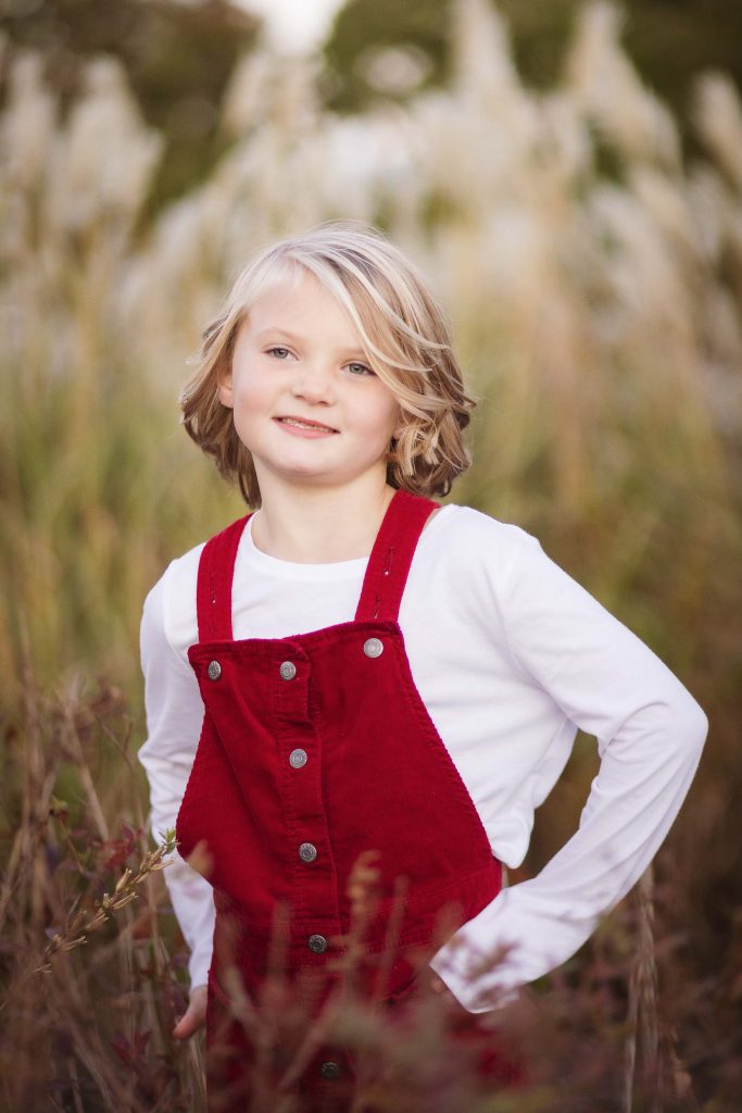 A little girl in red overalls standing in tall grass during sunset.