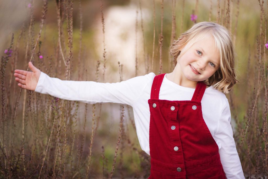 A little girl in red overalls standing in a field during sunset.