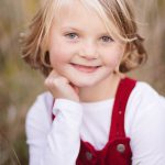 A little girl in a red overall sitting in tall grass during sunset.