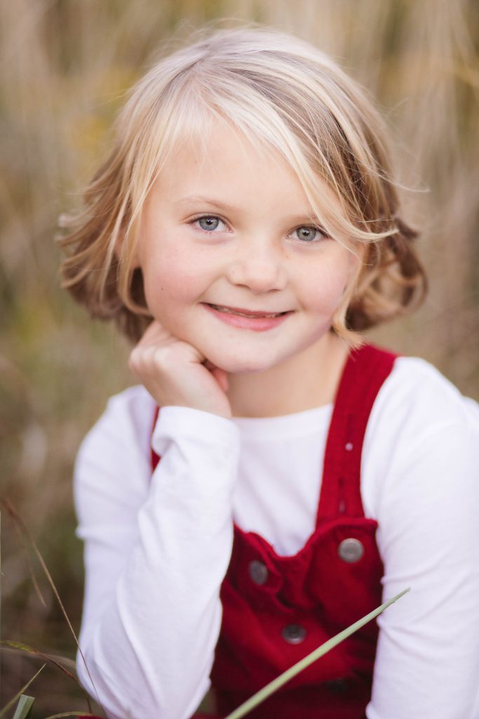 A little girl in a red overall sitting in tall grass during sunset.