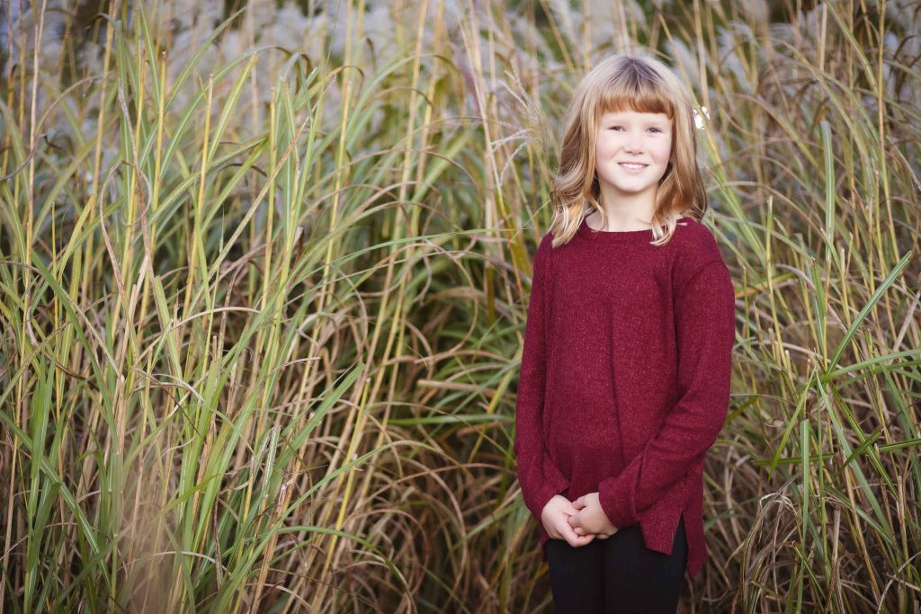 A little girl in a maroon sweater standing in tall grass during sunset.