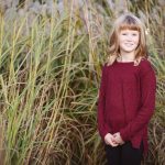 A little girl in a maroon sweater standing in tall grass during sunset.