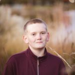 A boy in a maroon sweater standing in front of a lake during sunset.