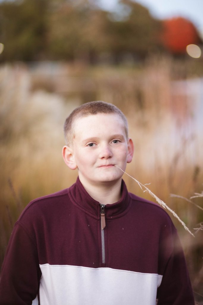A boy in a maroon sweater standing in front of a lake during sunset.