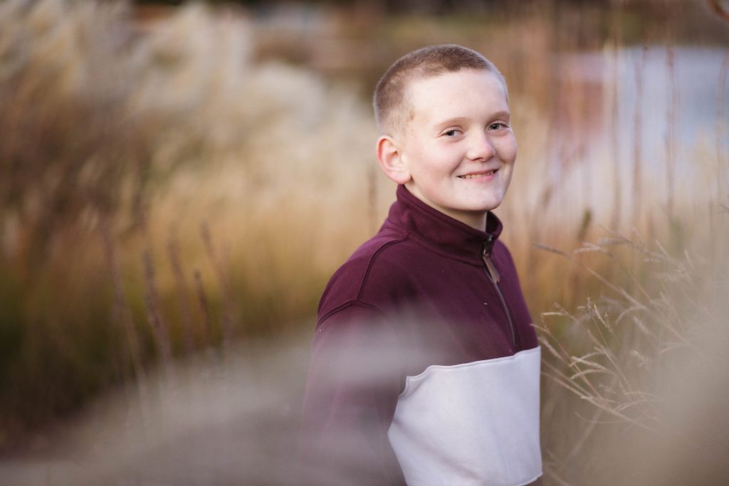 A boy smiles while standing in tall grass at sunset.