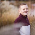 A boy smiles while standing in tall grass at sunset.