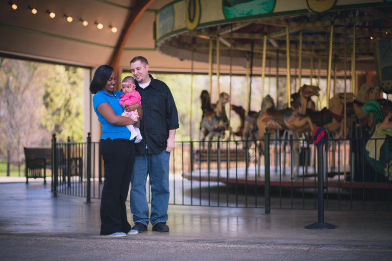 A couple with a baby are in Wheaton Regional Park, Maryland, posing in front of a carousel.