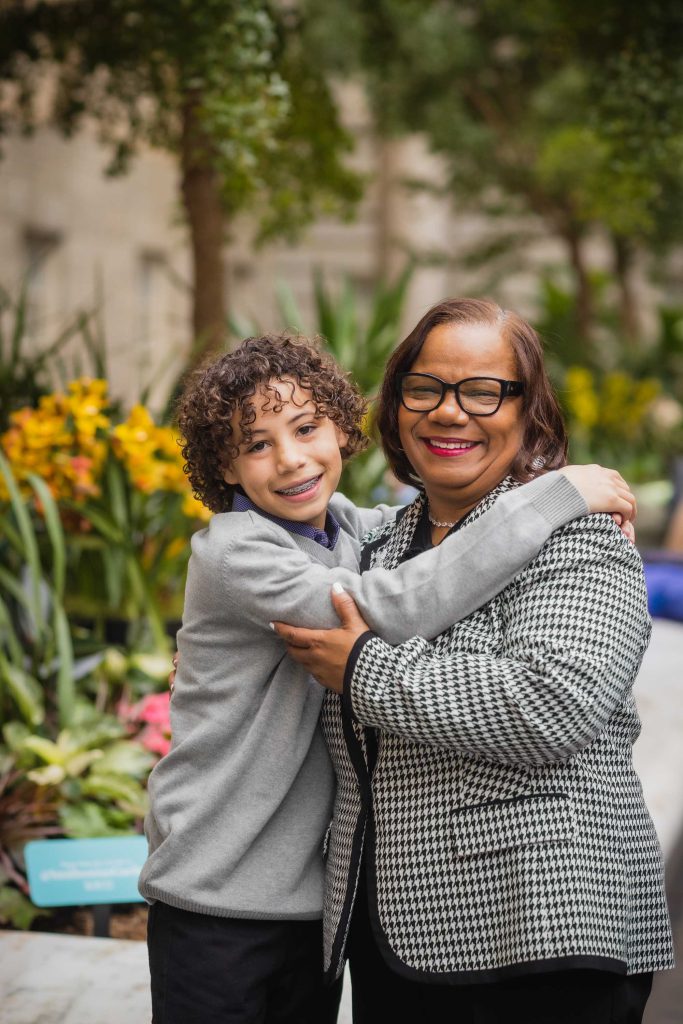 A family, consisting of a woman wearing glasses and a young boy, embraces lovingly in front of a flower garden.
