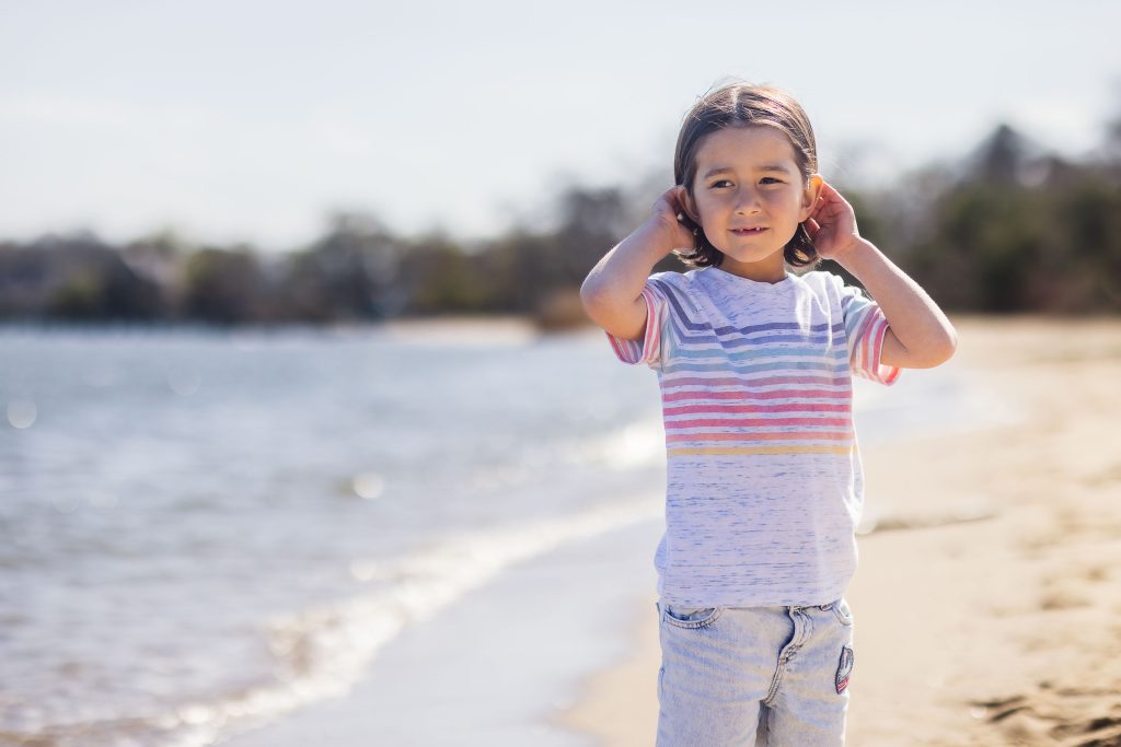 A portrait of a little girl standing on the beach with her hands on her ears.
