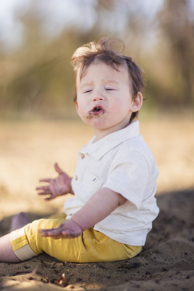 A portrait of a baby sitting in the sand in Annapolis, Maryland, with his mouth open.