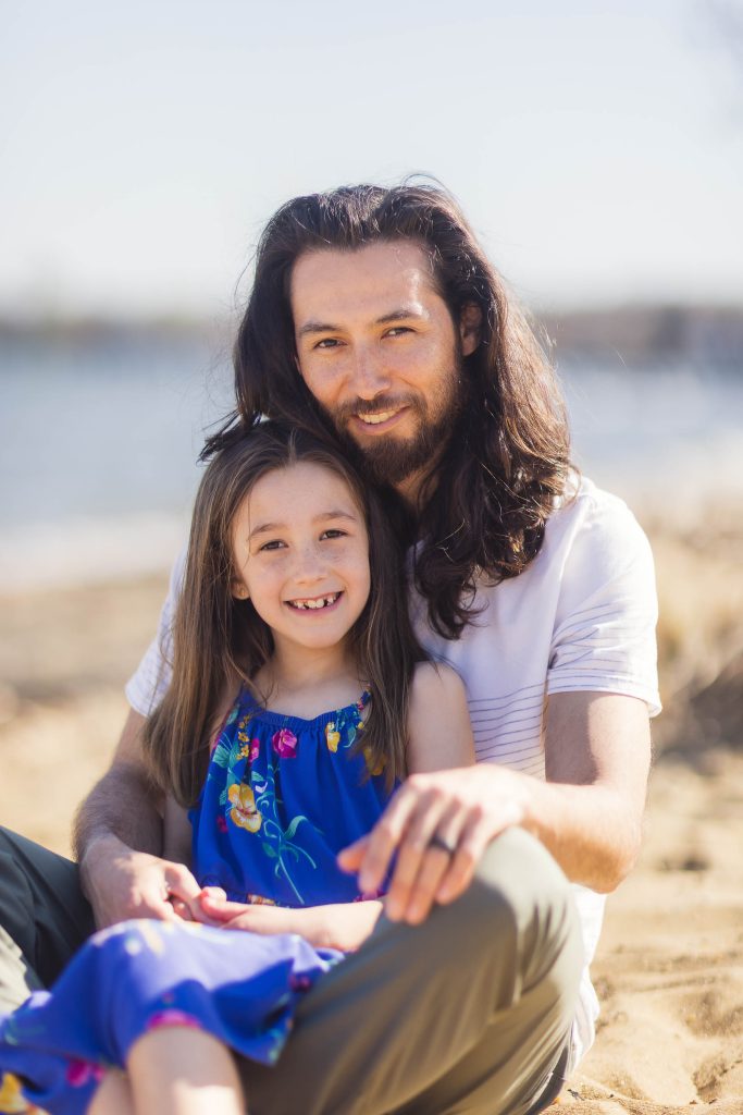 A man with long hair and a little girl sitting on the beach in Maryland, captured in a heartwarming portrait by the Chesapeake Bay Foundation.