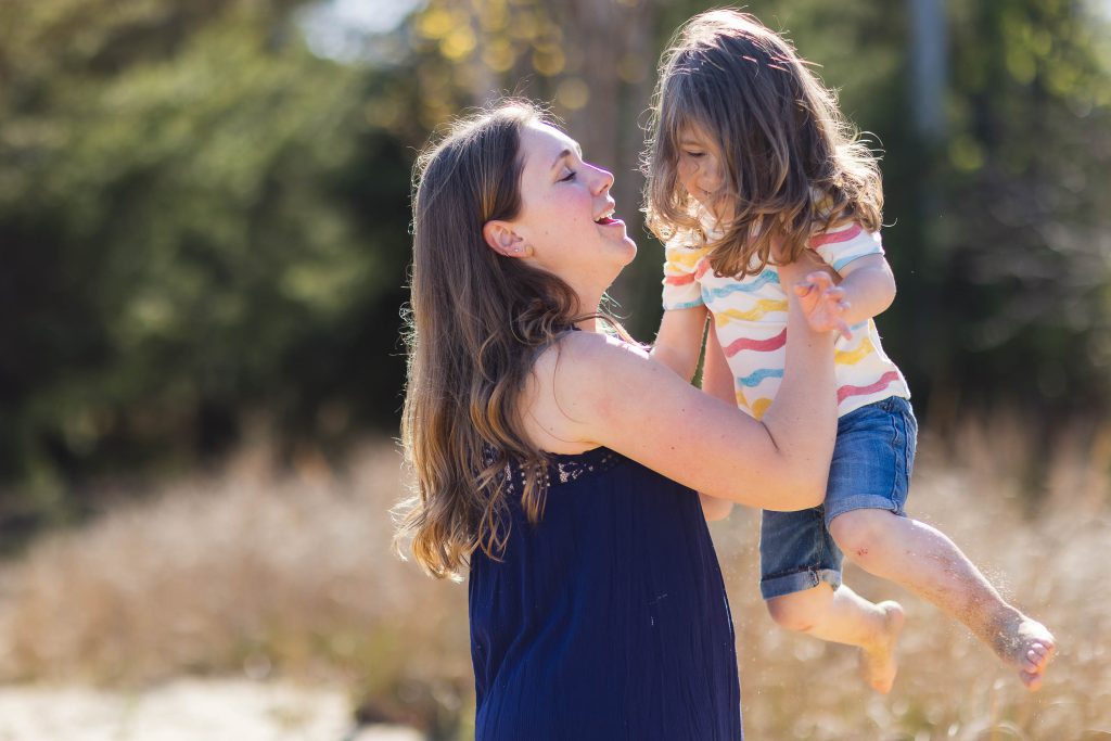 A mother is holding her daughter in the air in a field near Chesapeake Bay Foundation.