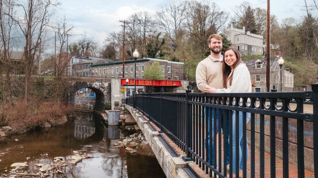 An engagement photo of a couple standing on a bridge over the Old Ellicott City river.