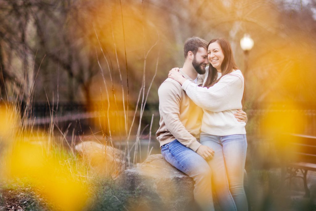 An engaged couple sits on a bench in Old Ellicott City, with yellow flowers in the background, capturing a beautiful portrait for their engagement photo.
