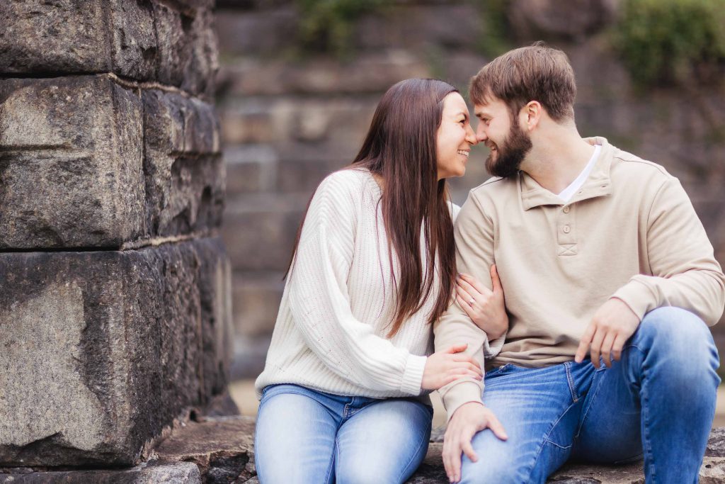 An engaged couple sits on a stone wall in Old Ellicott City, sharing a tender kiss as their portrait is captured for their engagement photo.