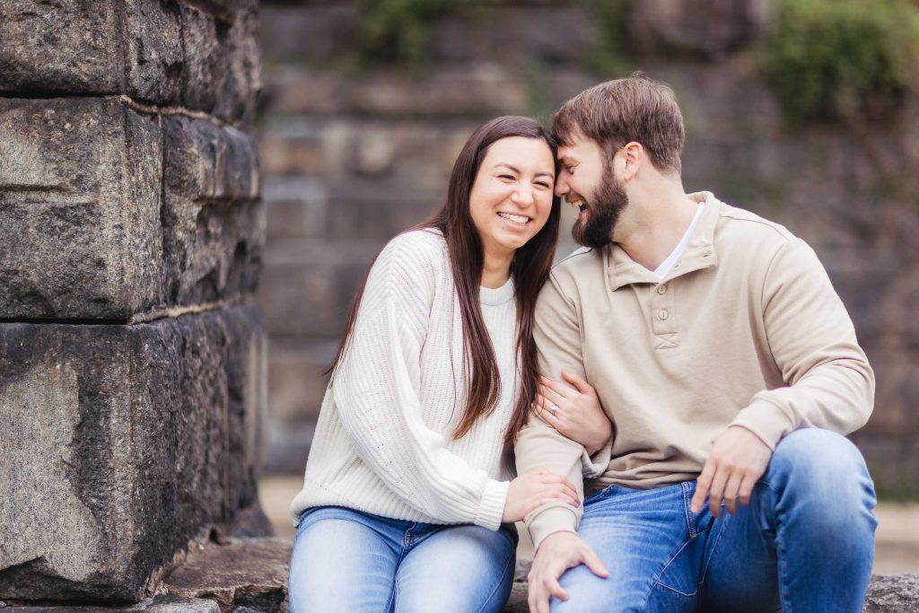 A couple poses for their engagement portrait in Old Ellicott City, sitting on a stone wall.