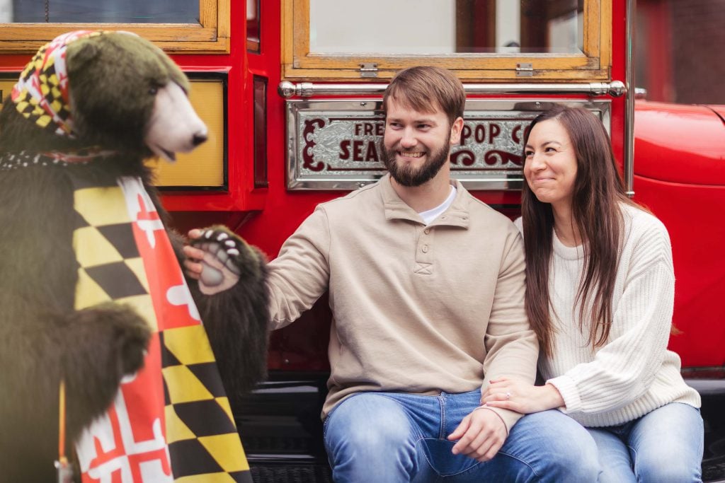 An engaged couple sits in front of a red trolley with a stuffed bear, capturing their love in a beautiful Old Ellicott City engagement photo.