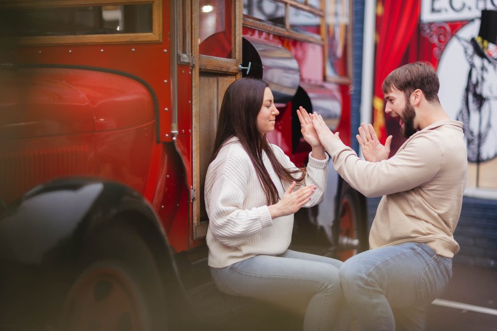 A couple is kneeling in front of a red bus, capturing an engagement portrait in Old Ellicott City.