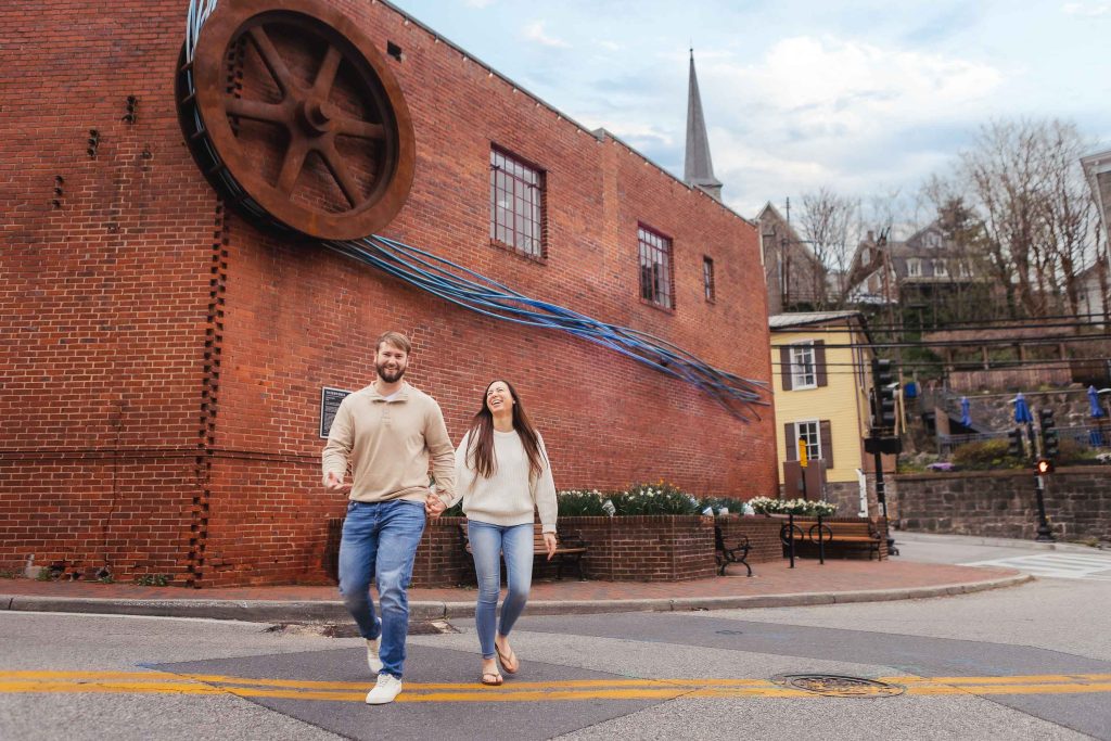 An engaged couple strolling down the street in front of an old red brick building, capturing a timeless portrait amidst the historic charm of Old Ellicott City.