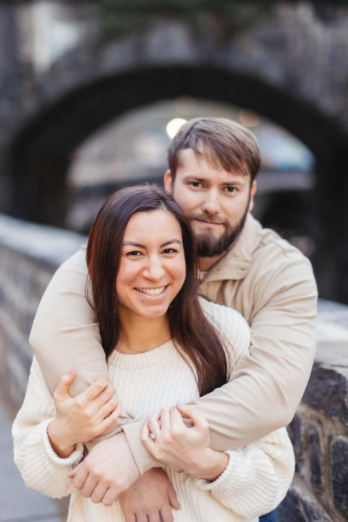 An engaged couple hugging in front of an old stone wall.