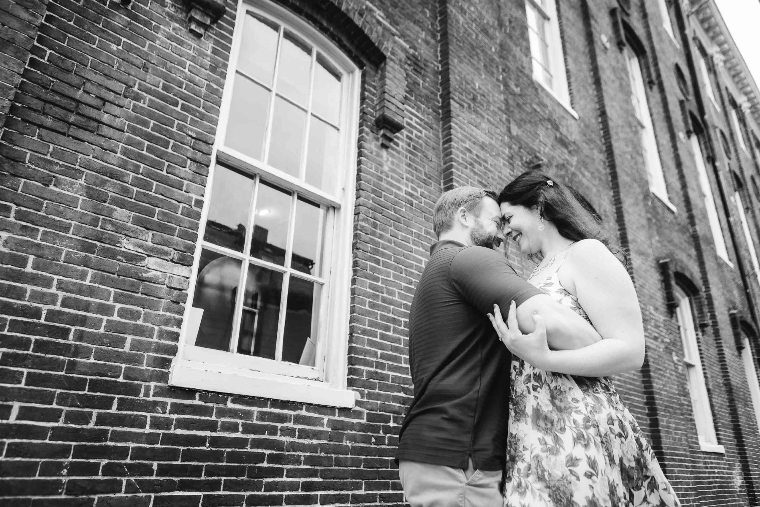 A couple embraces in front of a downtown Annapolis brick building while capturing their engagement photos.