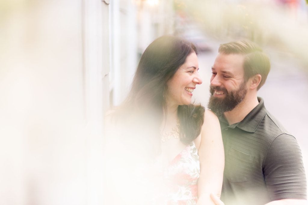 A couple is smiling while leaning against a wall in downtown Annapolis for engagement photos.