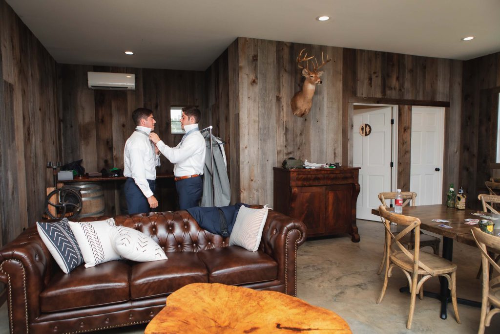 Two men, dressed in wedding attire, candidly putting on their ties in a living room at the Fleetwood Farm Winery.