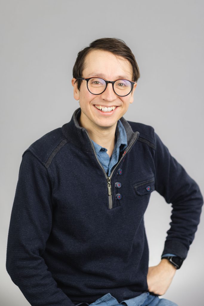 A smiling man in glasses sitting on a gray background.